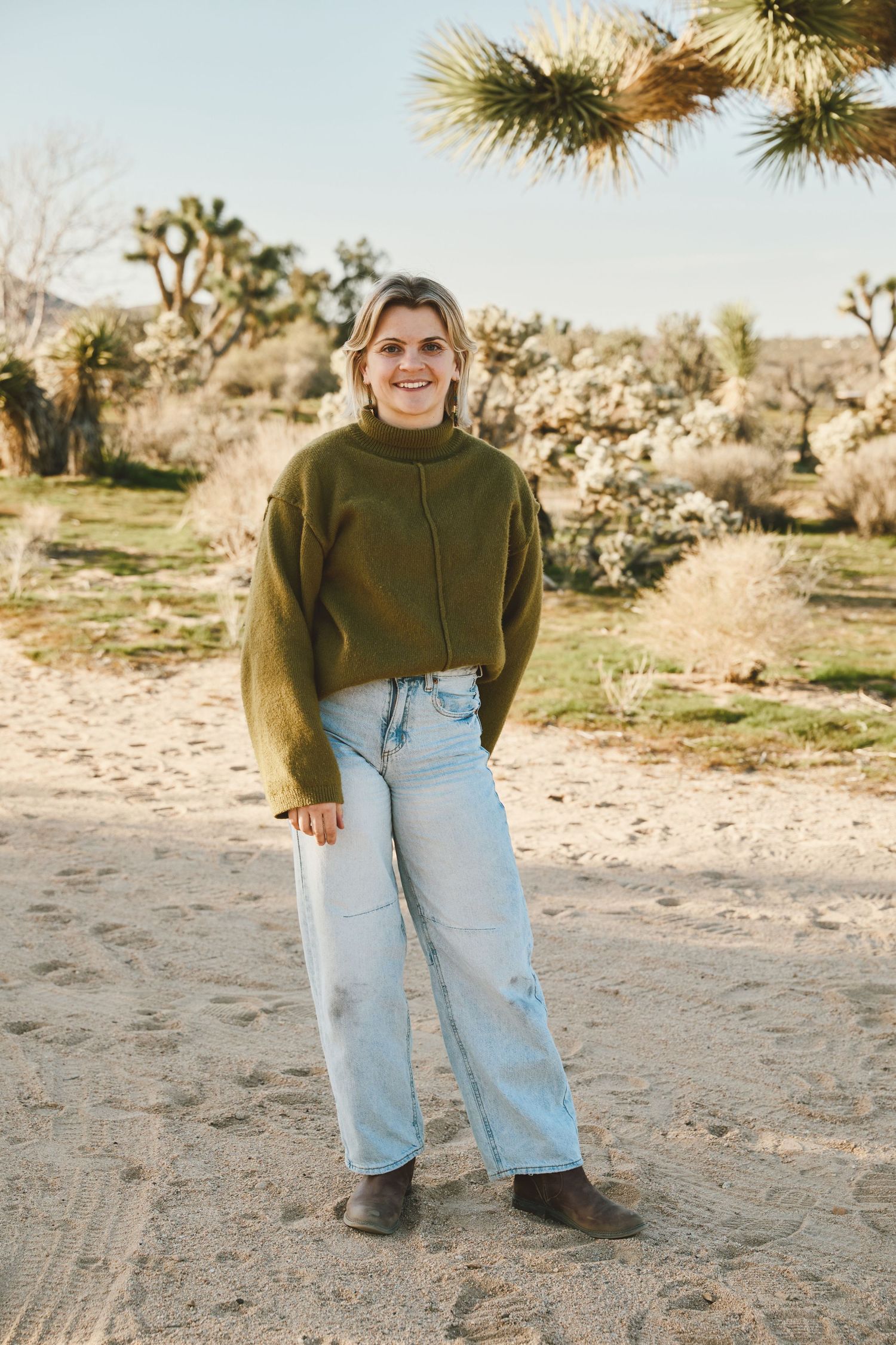 A person wearing an olive green sweater and light wash jeans stands in a desert landscape with Joshua trees in the background.