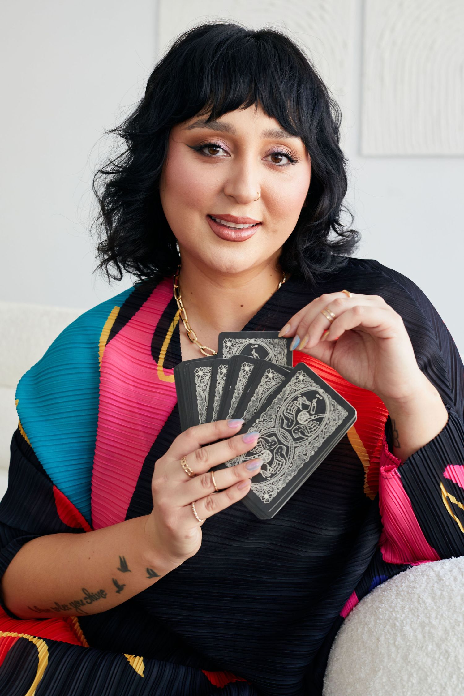 Woman holding dark tarot cards in colorful kimono-style top, black hair with bangs, gold jewelry, tattoos.