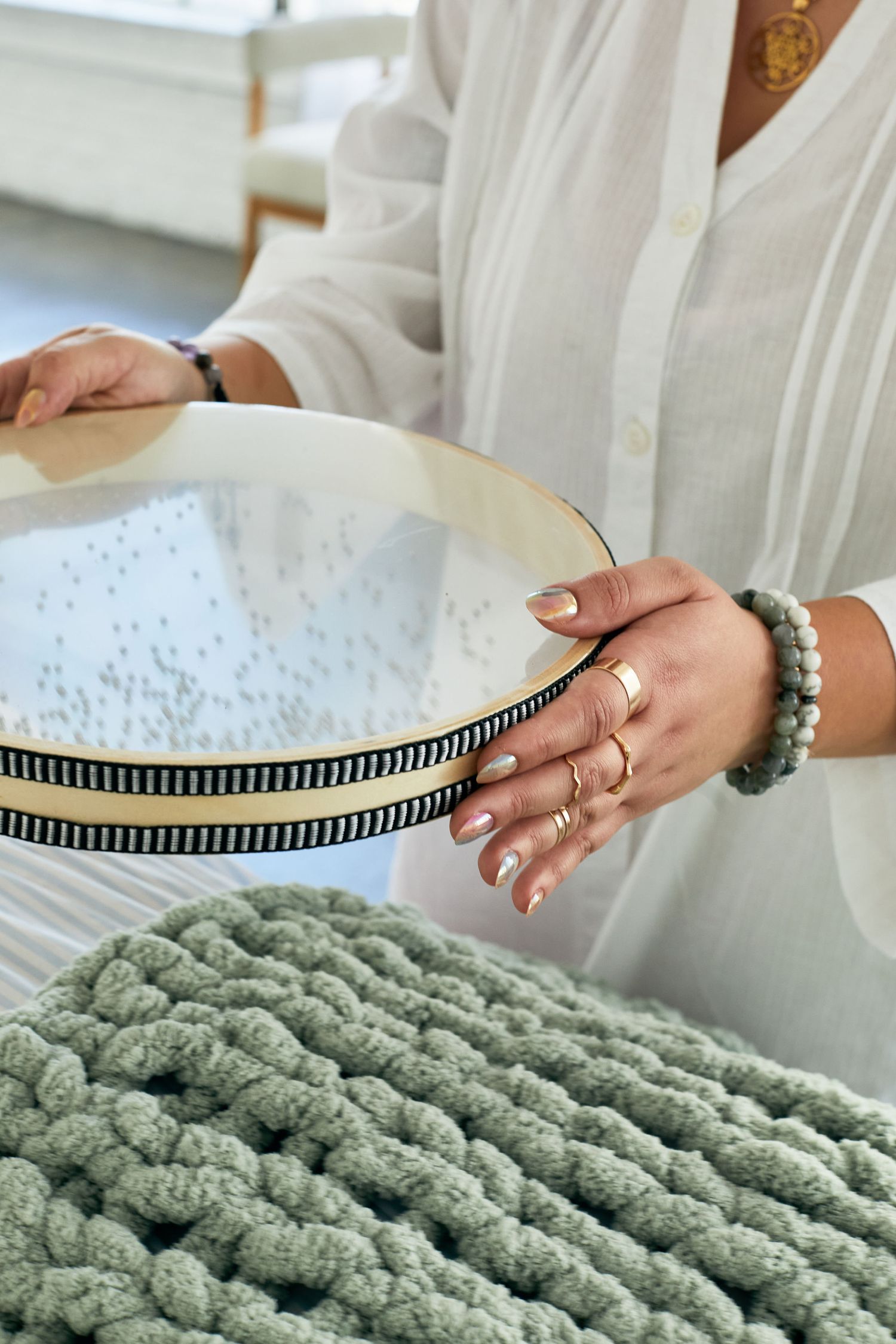 Woman in white shirt holding embroidery hoop with rings and bracelet, sage green chunky knit blanket in foreground.