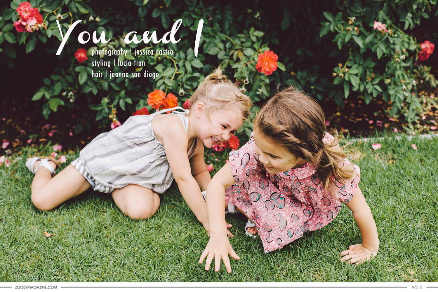 Two children playing and laughing together on a green lawn with flowering bushes in the background.