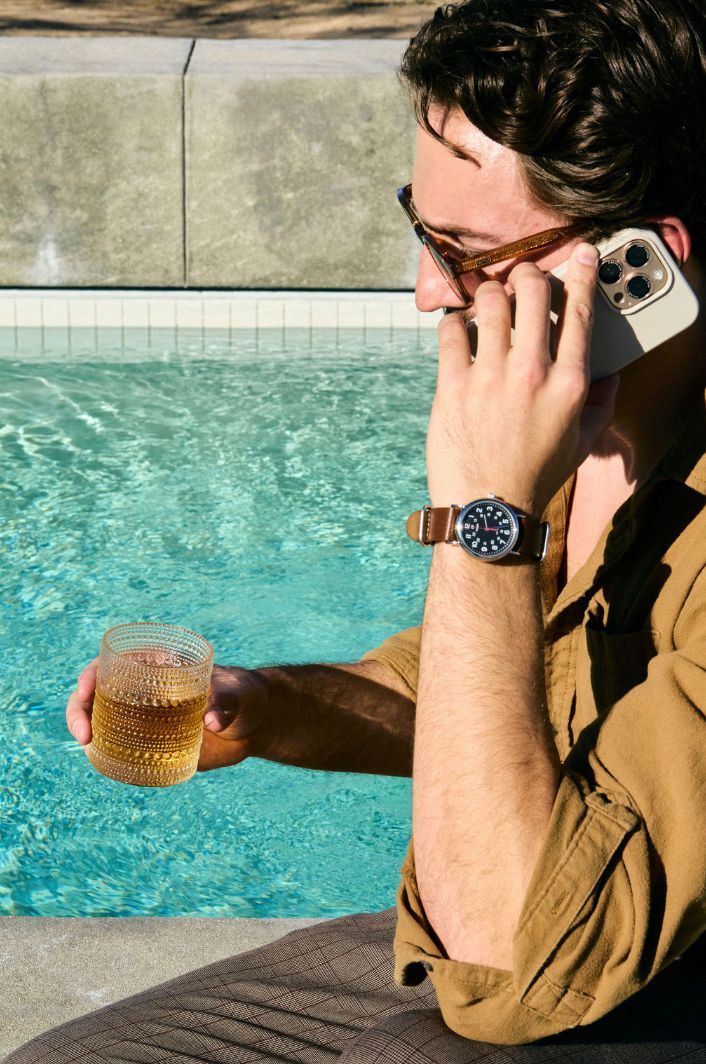 A person in a brown jacket holds a glass of whiskey while sitting by a swimming pool on a sunny day.