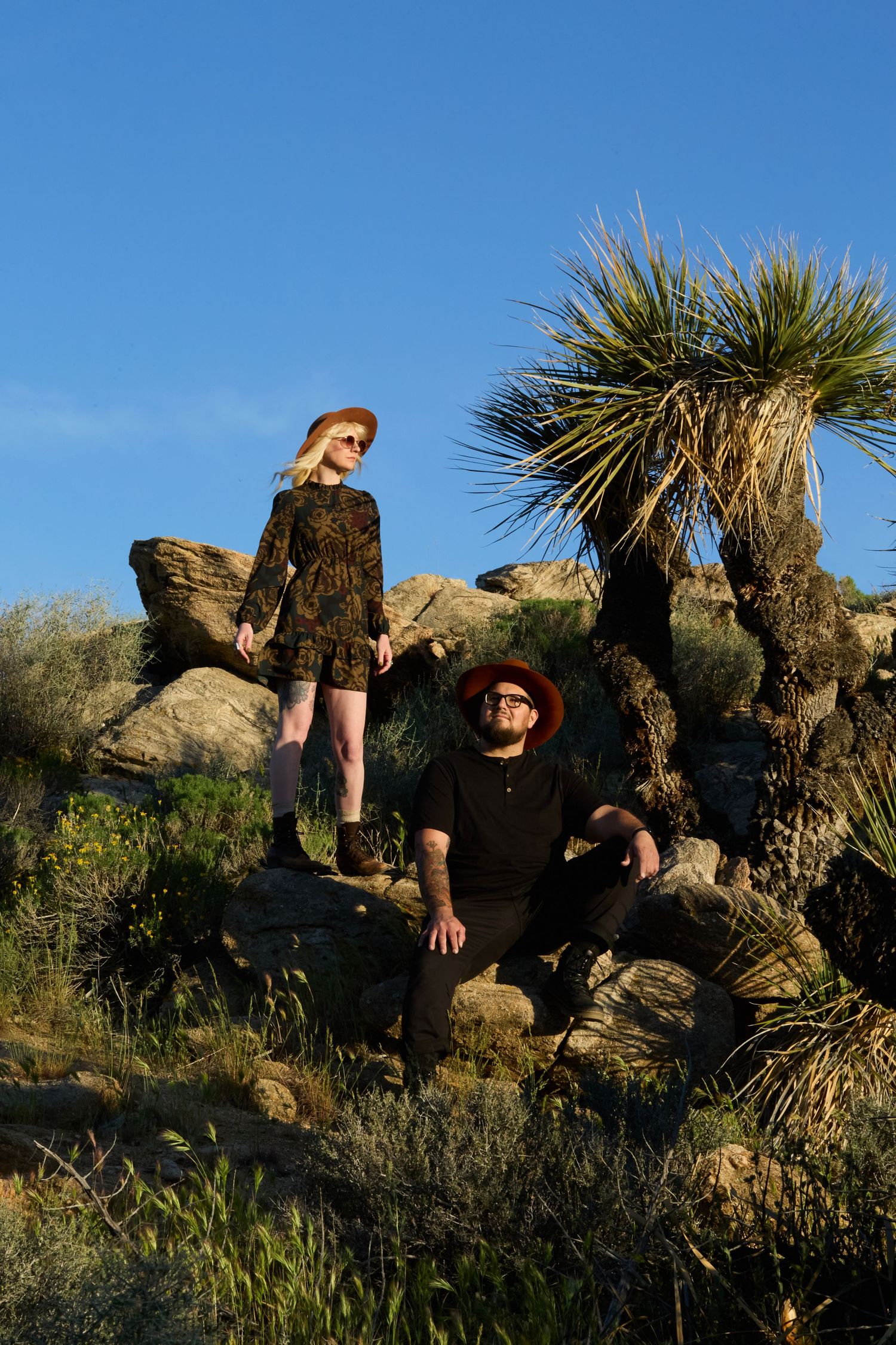 A woman in a floral dress and man in black sit among desert rocks and Joshua trees under a clear blue sky.