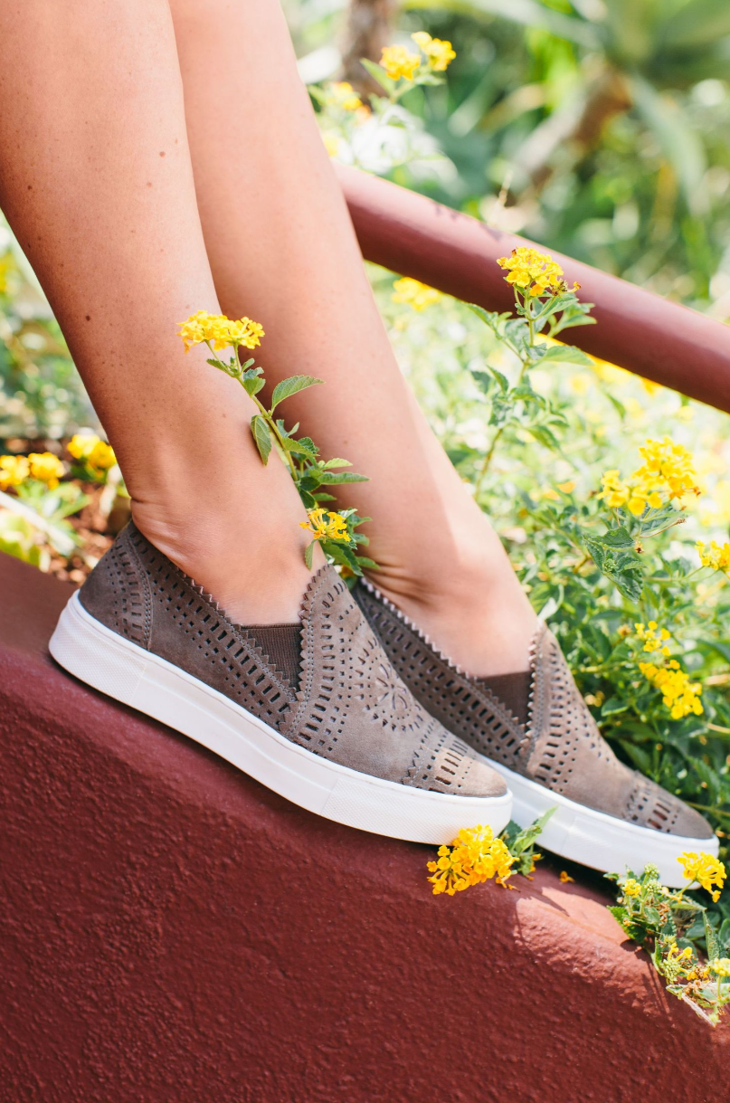 Grey perforated slip-on sneakers surrounded by yellow flowers.