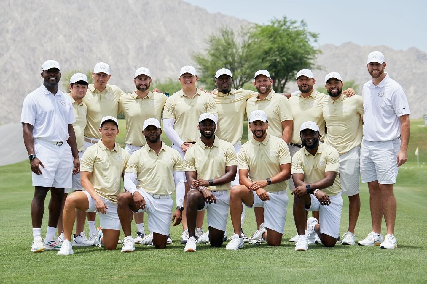 Tennis team members in white and light yellow uniforms pose together for a group photo on an outdoor court with mountains visible.