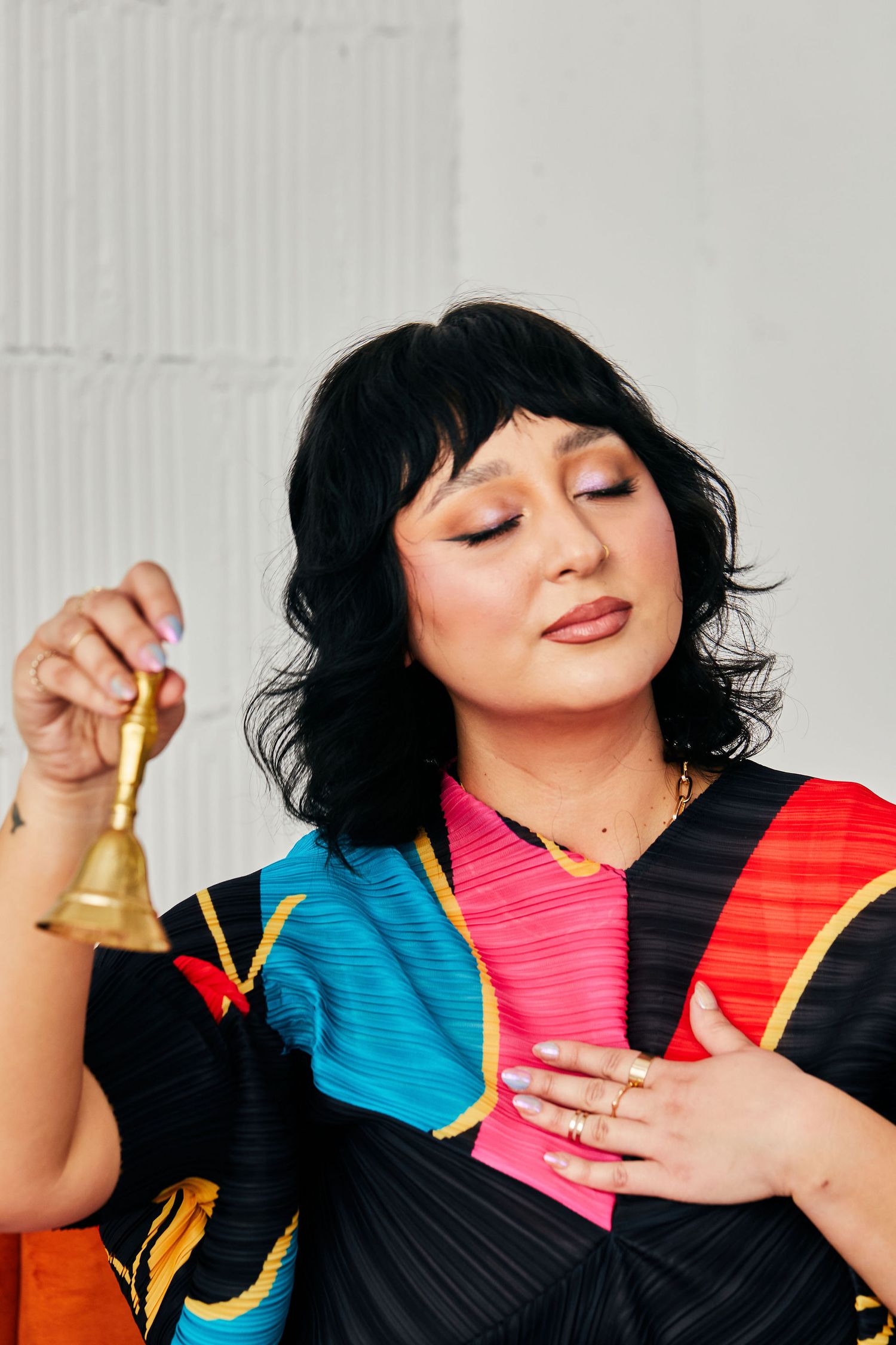 Woman in colorful black, pink, and blue outfit holding a brass bell with eyes closed and hand on chest.
