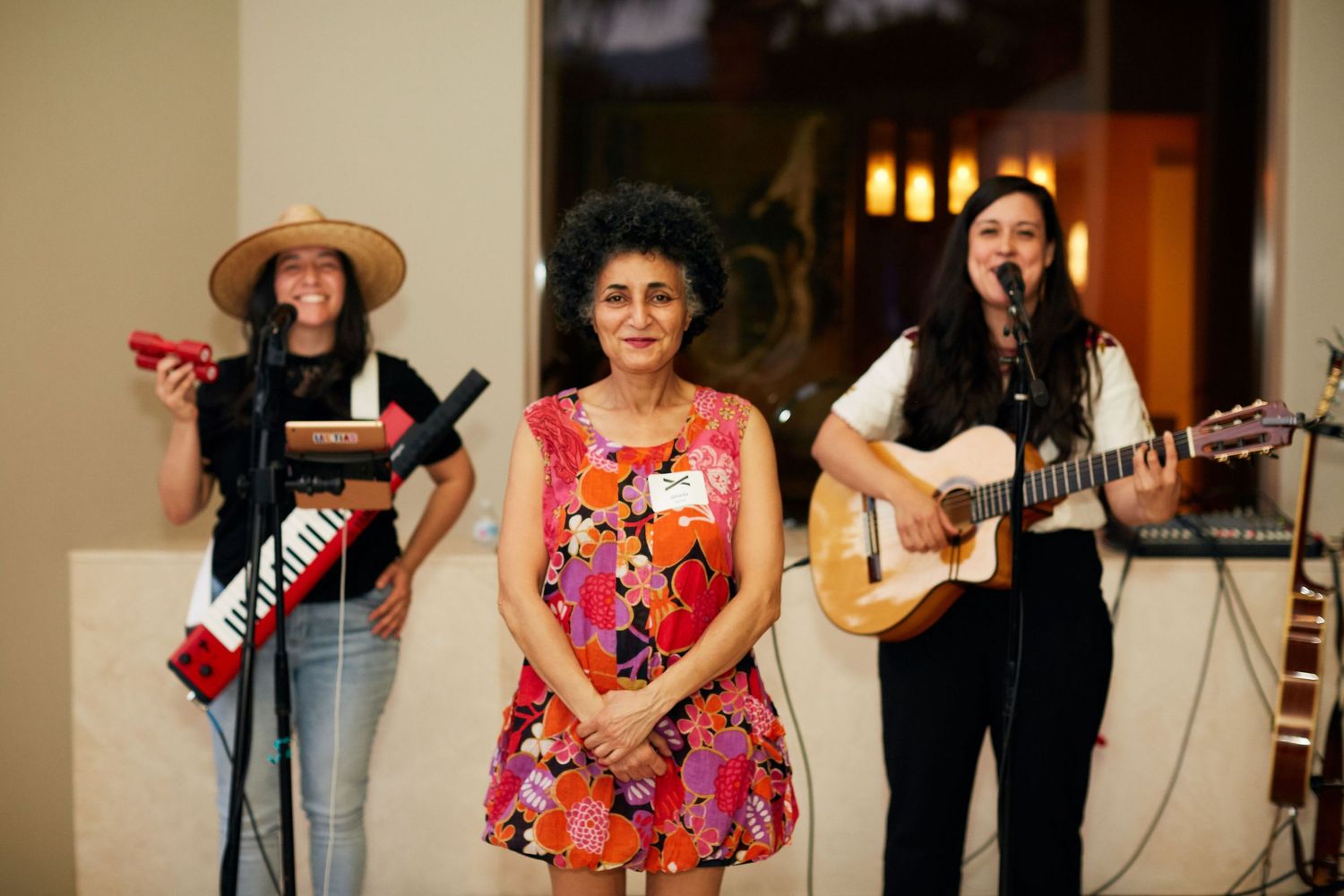 Three musicians perform together, with two playing guitars while wearing casual attire in a warmly lit indoor setting.