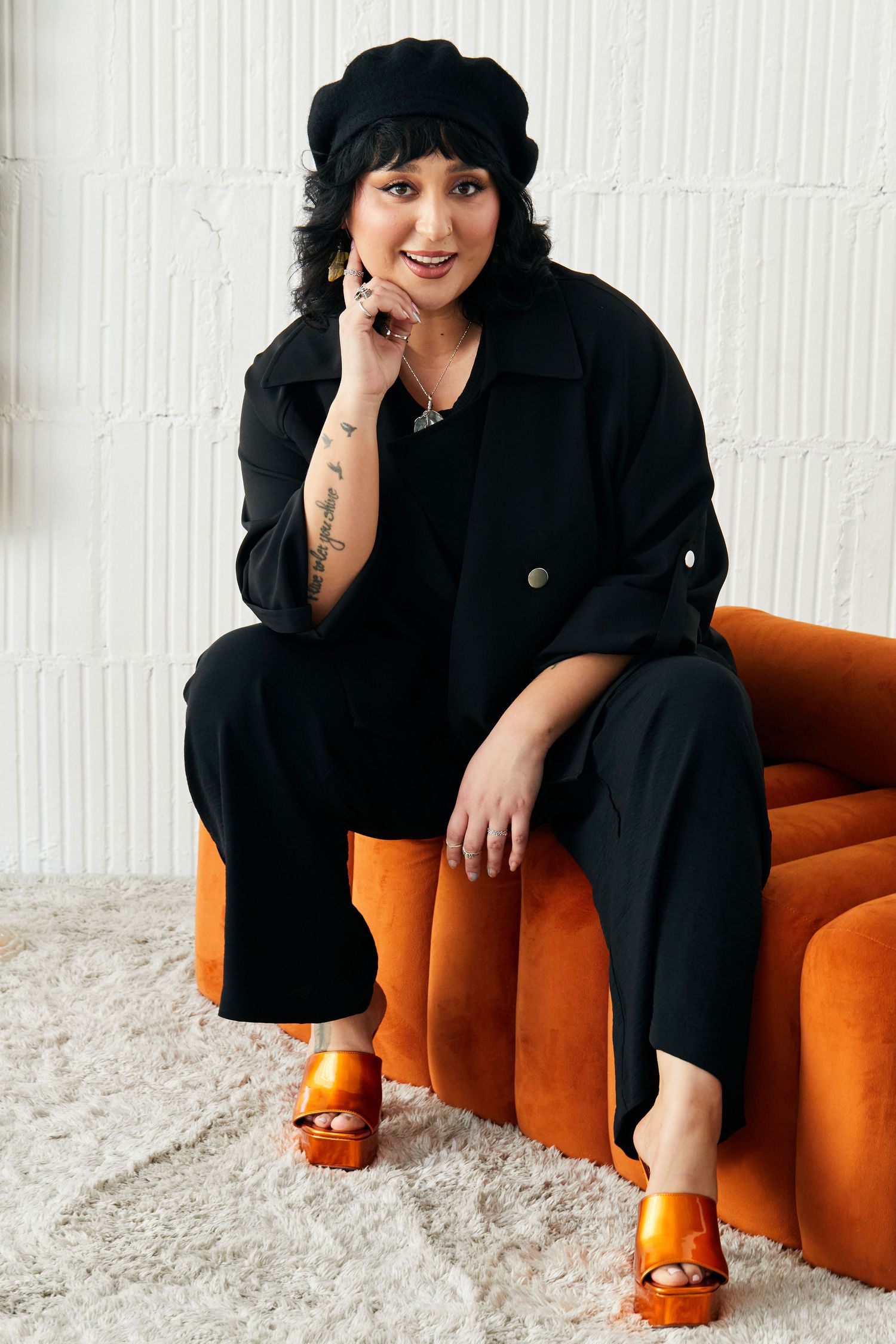 Woman in black outfit and beret sits on orange velvet chair, wearing orange platform sandals, smiling confidently.
