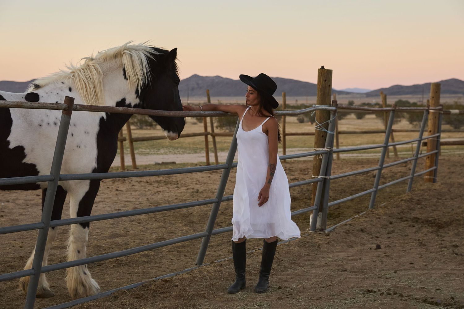 Woman in white dress and cowboy hat standing beside a paint horse at a fence during sunset.