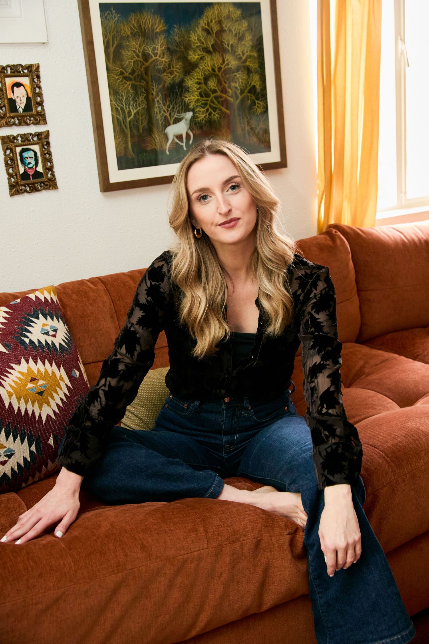 Blonde woman in black floral top and jeans sitting on an orange velvet sofa with artwork on the wall.