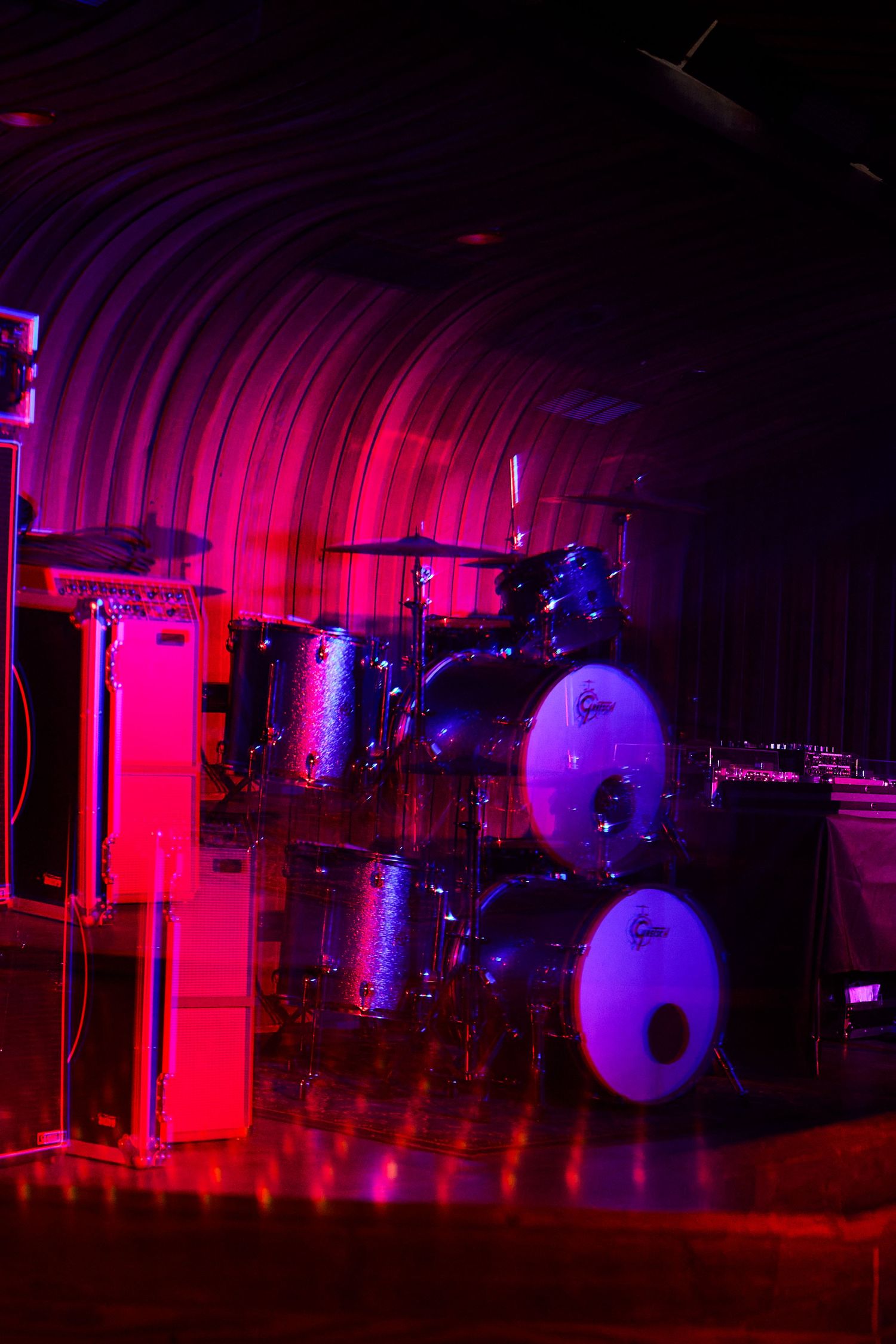 A drum kit illuminated by vibrant pink and purple stage lighting creating a dramatic nightclub atmosphere.