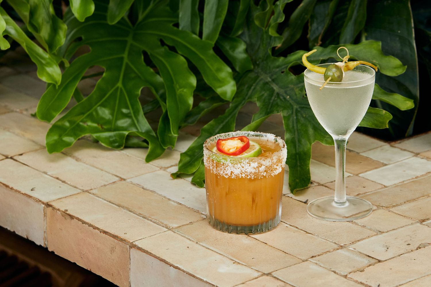 Cocktail and orange drink garnished with citrus on stone patio near tropical plants.