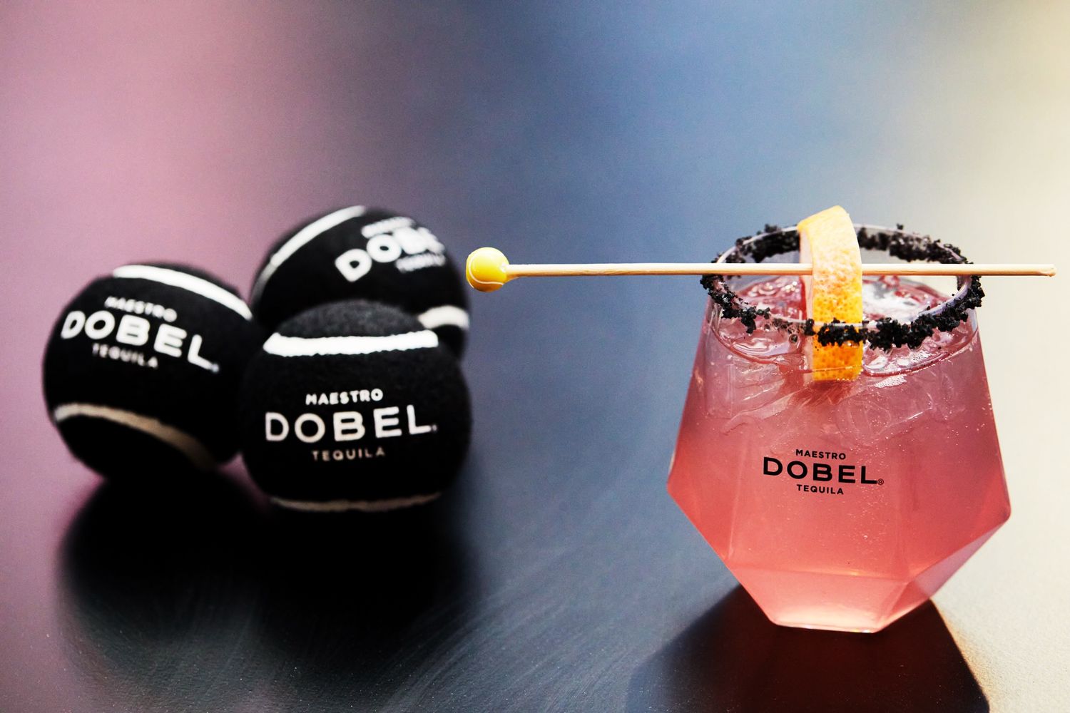 Pink DOBEL tequila cocktail with black branded accessories on dark surface.