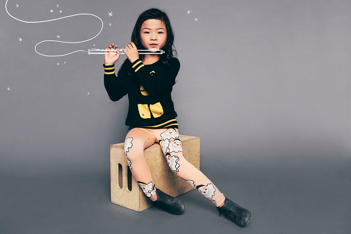 A musician in a black crop top and patterned tights holds a flute while sitting on a wooden box against a gray background.
