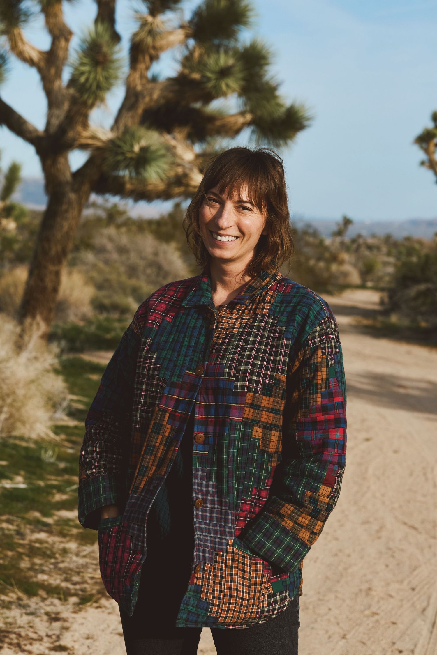 Someone wearing a colorful patchwork jacket stands on a desert trail with Joshua trees in the background.