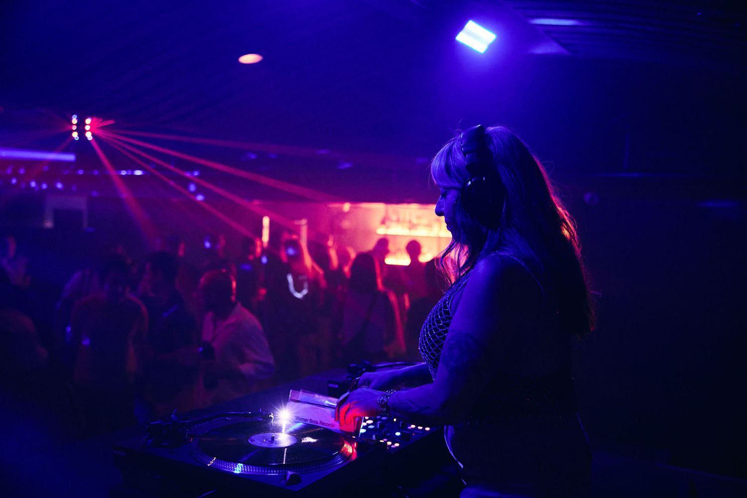 DJ performing at a nightclub with vibrant purple and red lighting effects illuminating the dance floor and stage area.