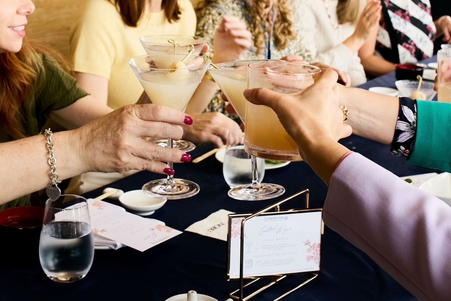 Close-up shot of hands holding martini glasses during a social gathering at a table with menu cards.
