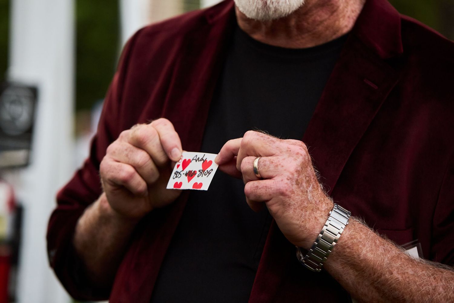 Close-up of hands holding a playing card while wearing a burgundy shirt and metal watch.