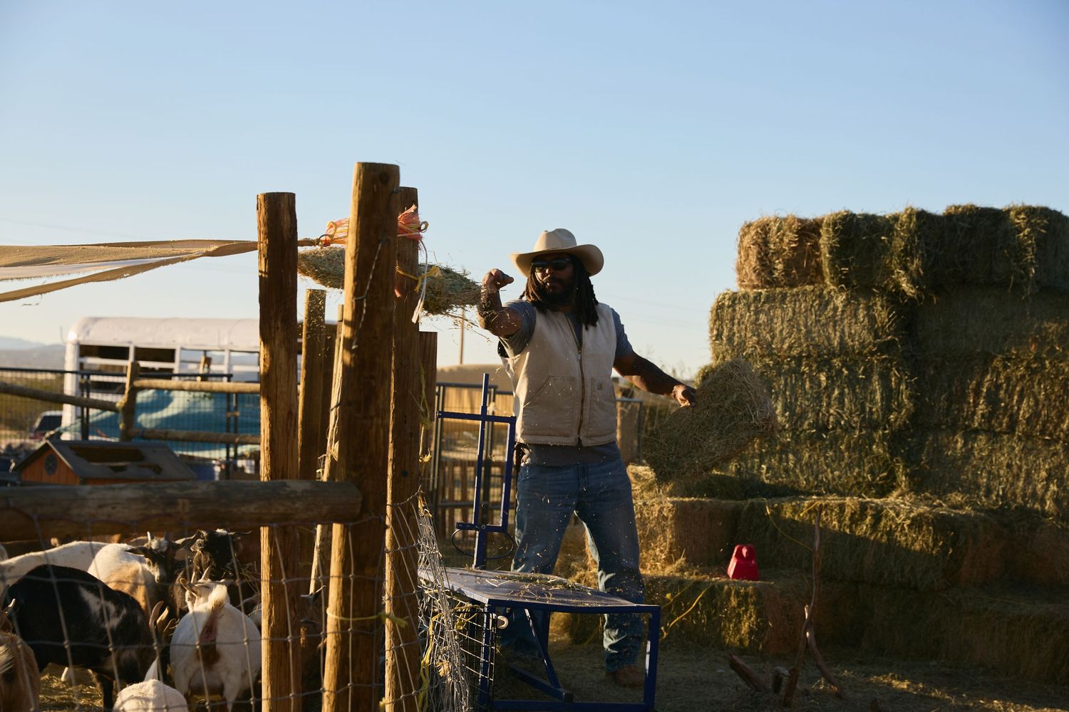 Farmer in cowboy hat working near wooden fence posts with stacked hay bales on a sunny rural farm.
