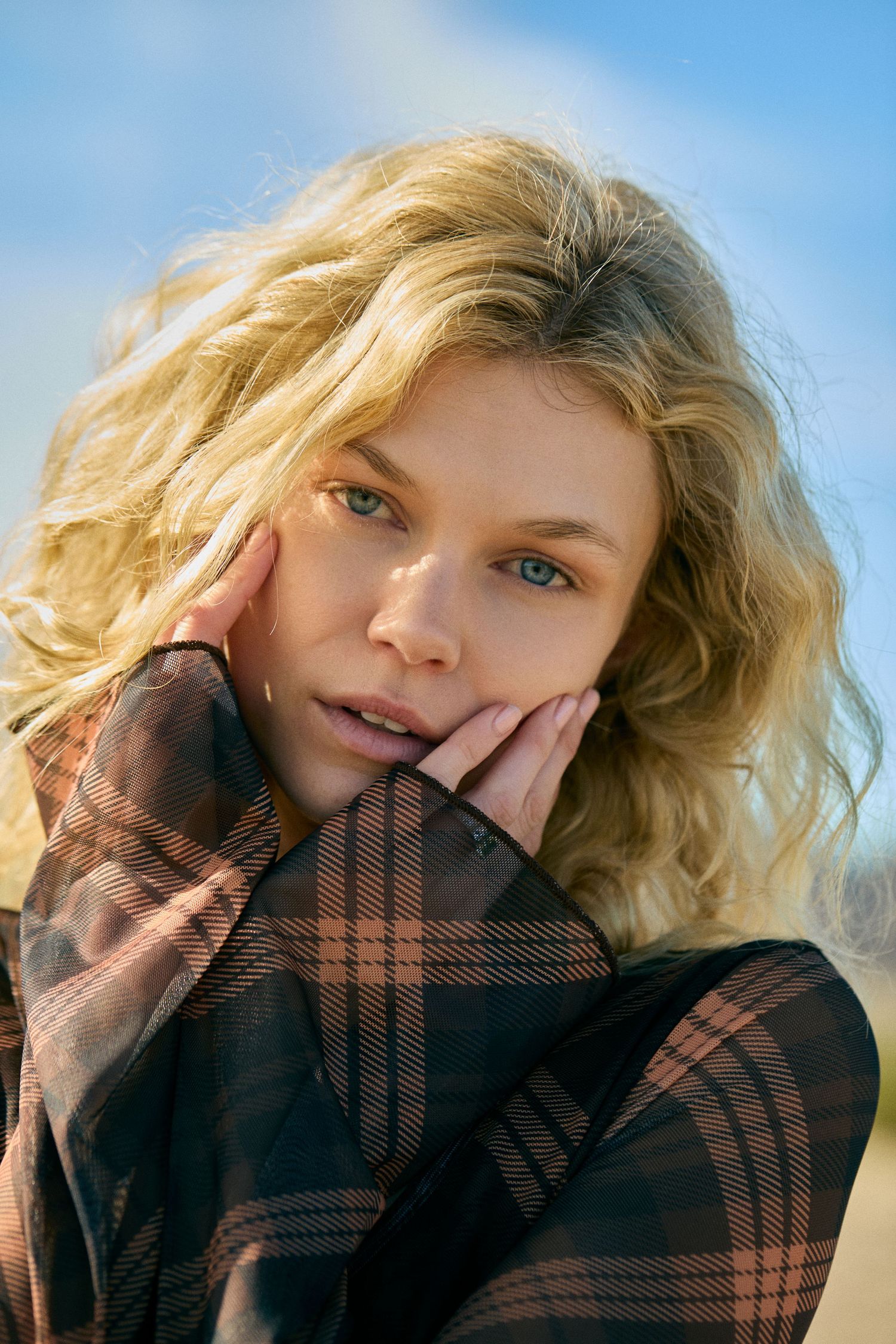Fashion portrait of blonde model wearing black mesh top against blue sky background.
