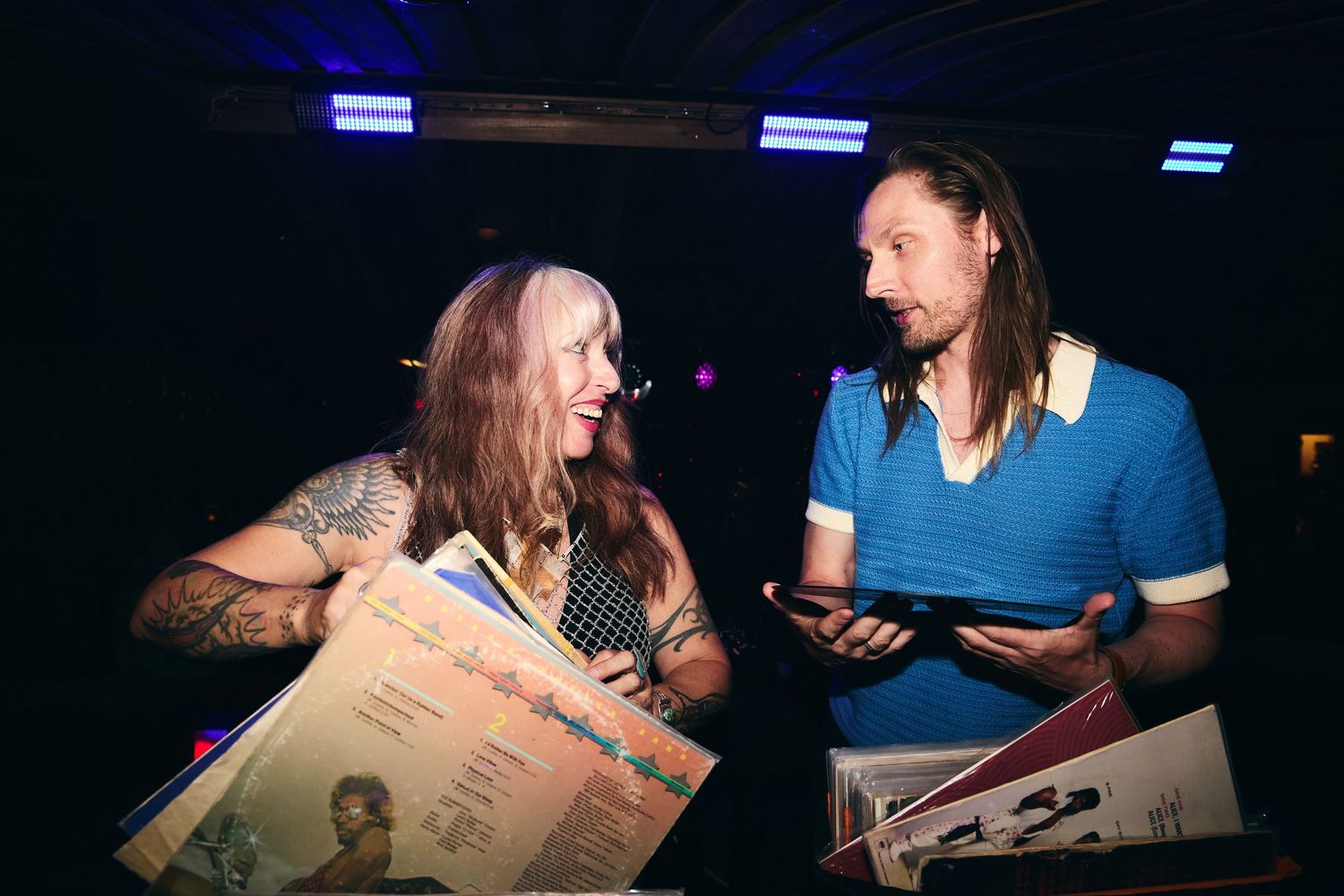 Two people engaging in conversation while holding vinyl records in a dimly lit space with blue lighting.