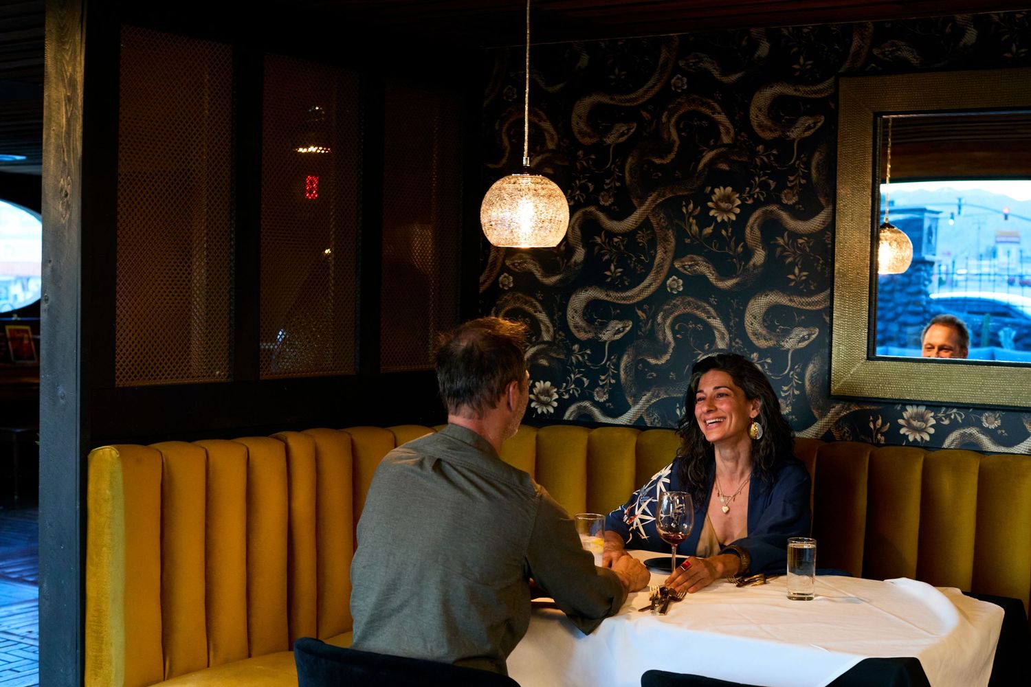 Diners enjoying an intimate conversation at a restaurant booth with yellow velvet seating and decorative wallpaper.