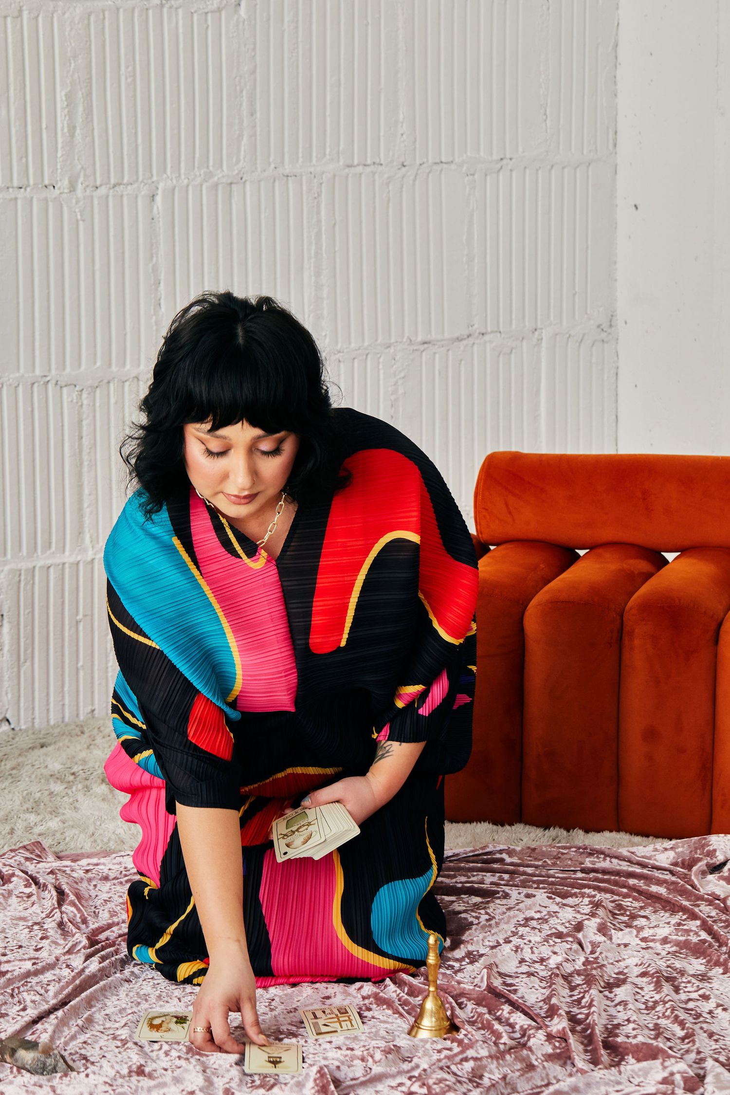 Woman in colorful black kimono kneeling on velvet fabric, holding tarot cards near a vintage orange sofa.