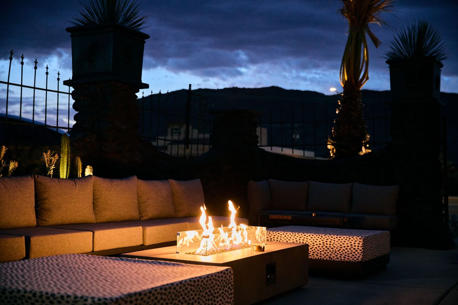 A modern outdoor lounge area with a linear fire pit illuminating cushioned seating at dusk against mountain silhouettes.