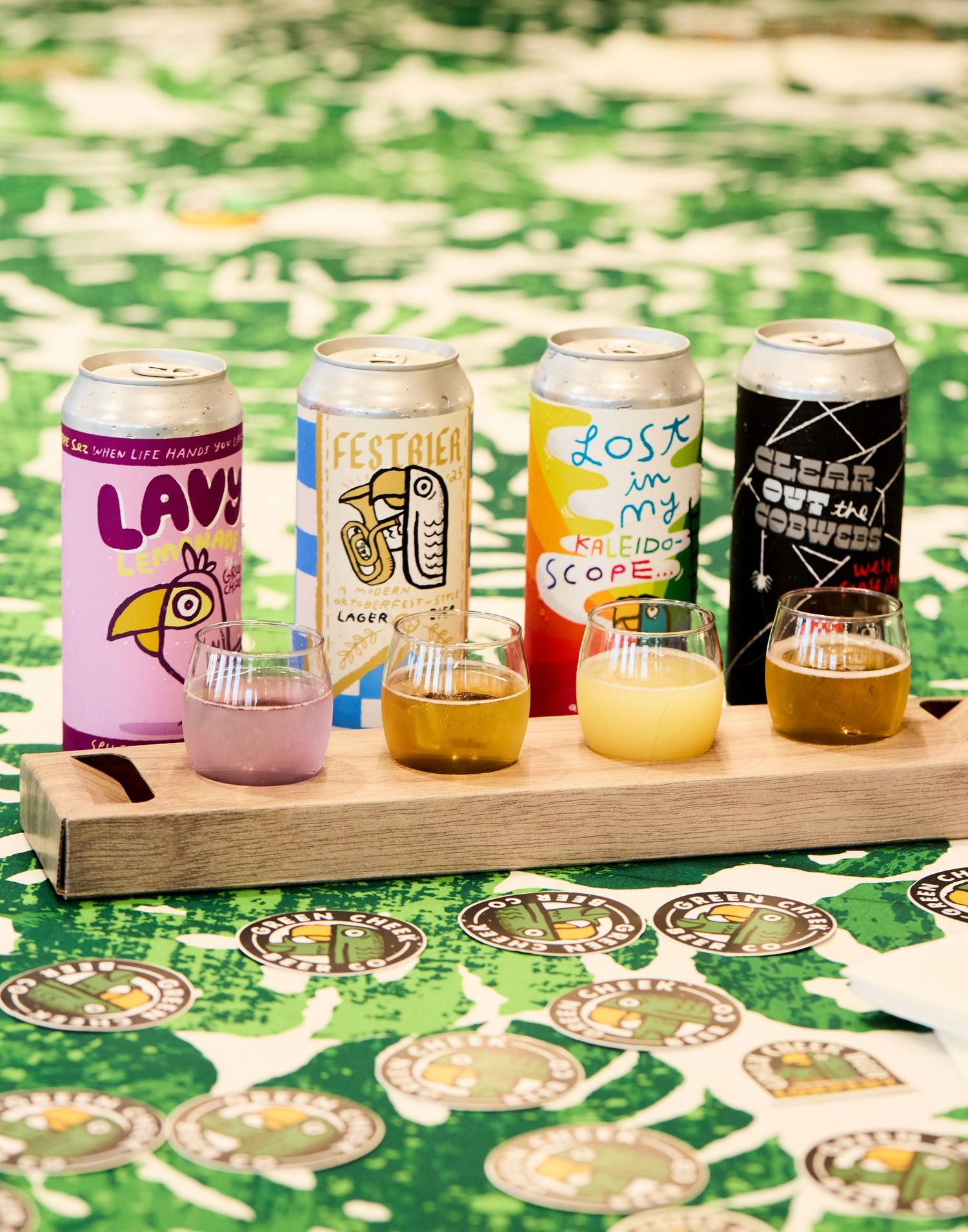 A wooden flight board displays four colorful craft beer cans with matching sample glasses on a patterned tablecloth.