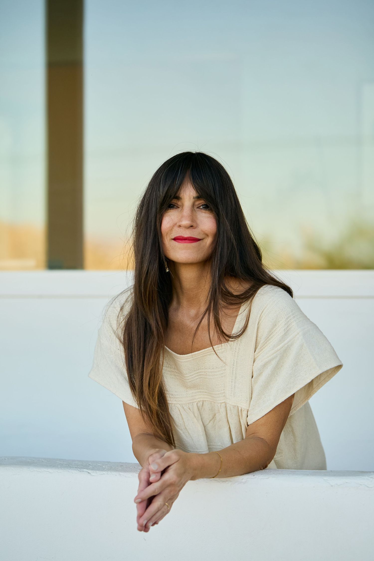 A person in a white top smiles while leaning on a sunlit white wall during a warm sunset.