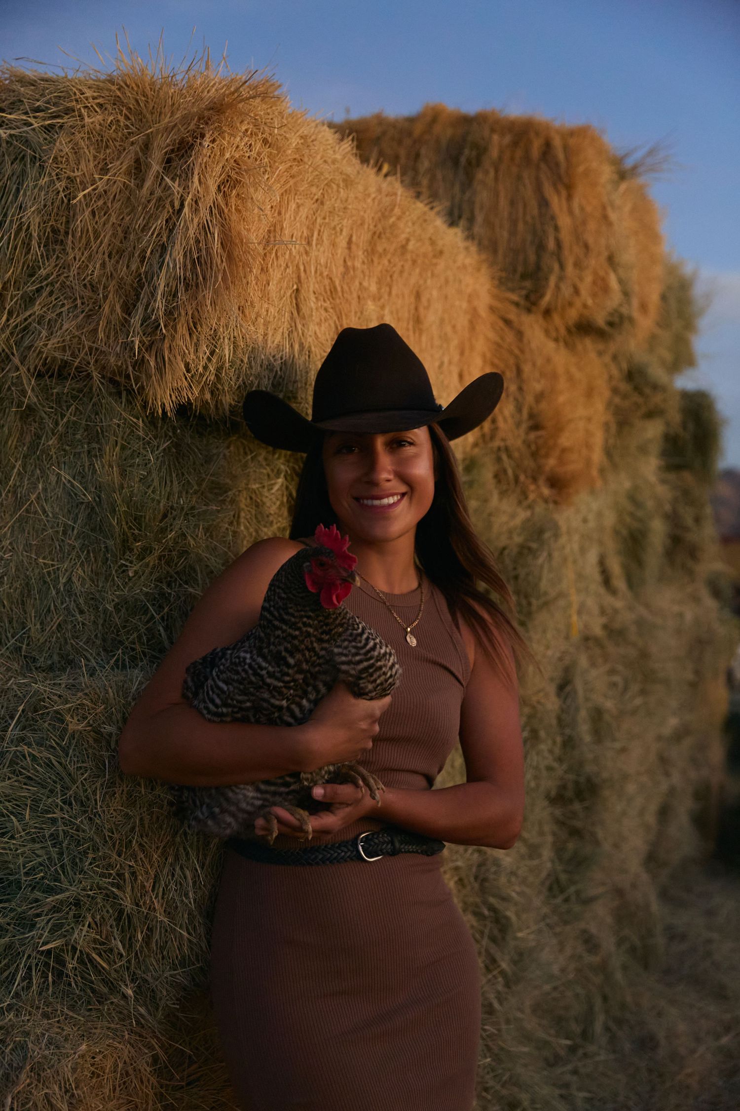 Smiling woman in black cowboy hat holding a chicken while leaning against hay bales at golden hour.