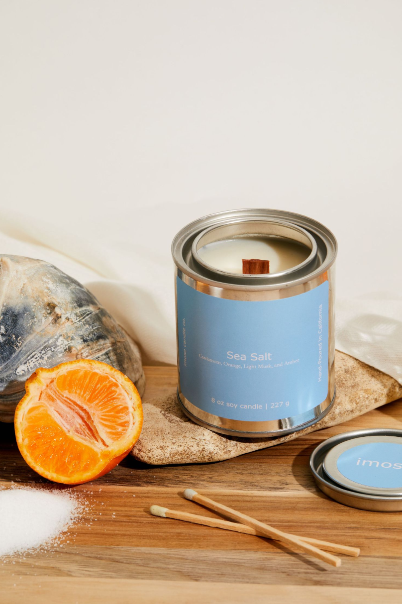 Blue candle tin displayed with orange slice and matchsticks on wooden surface.