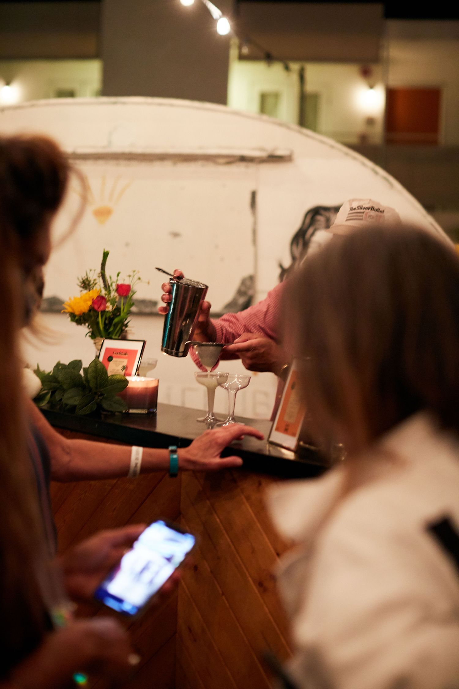 People gathered at a table with drinks, flowers and candles while one person checks their phone in dimly lit room.