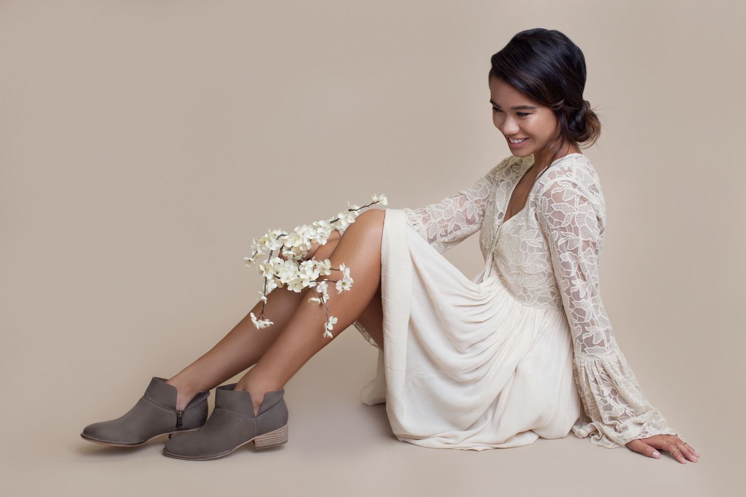 A bride poses in an elegant white lace wedding dress and gray ankle boots against a neutral background.