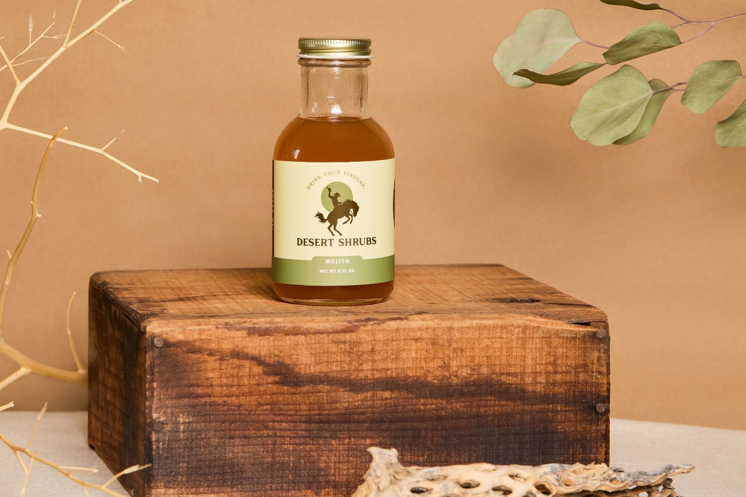 Desert Shrub herbal syrup in a glass bottle displayed on a rustic wooden block with eucalyptus branches.
