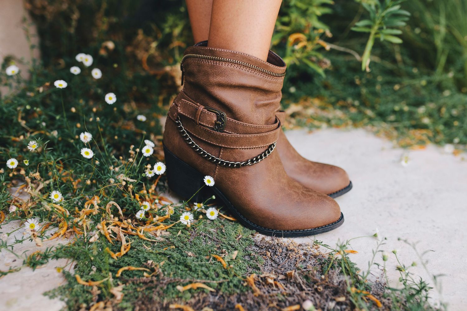 Brown leather ankle boots with chain detail photographed against grass and flowers.