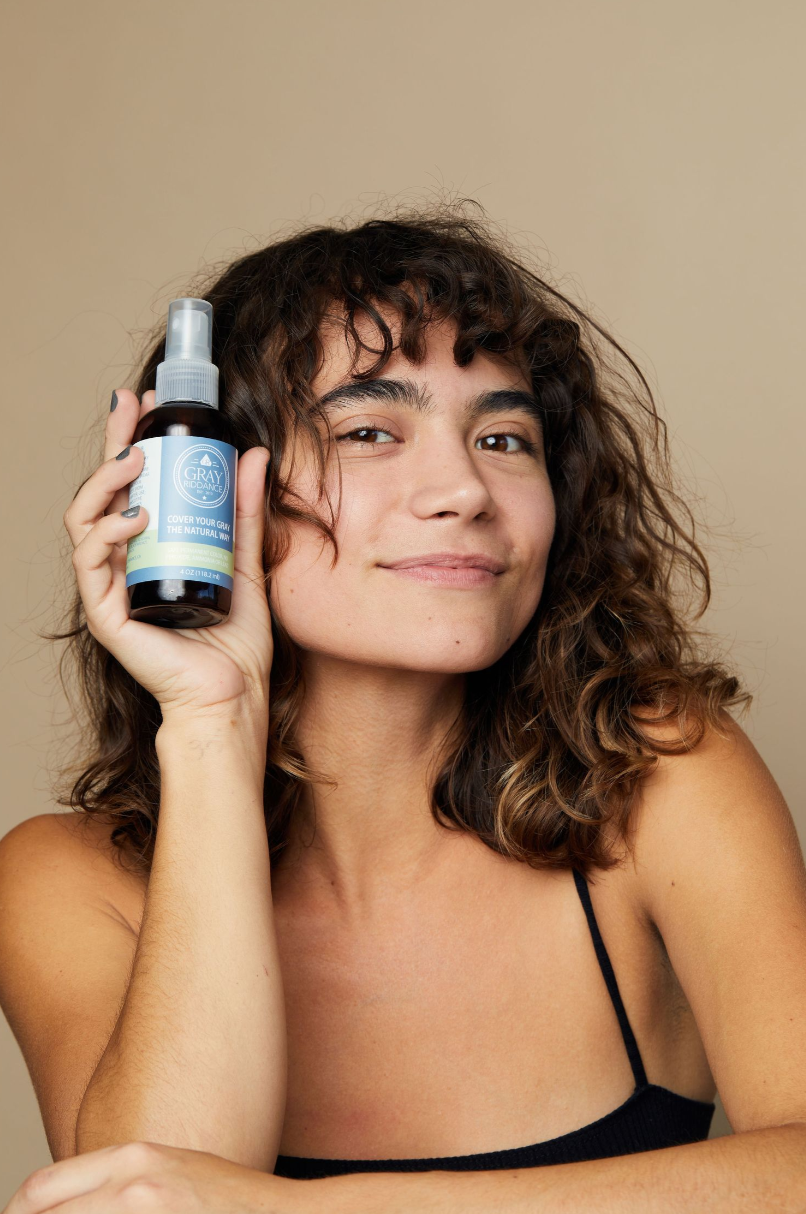 Beauty product advertisement showing spray bottle held against beige background in lifestyle shot.