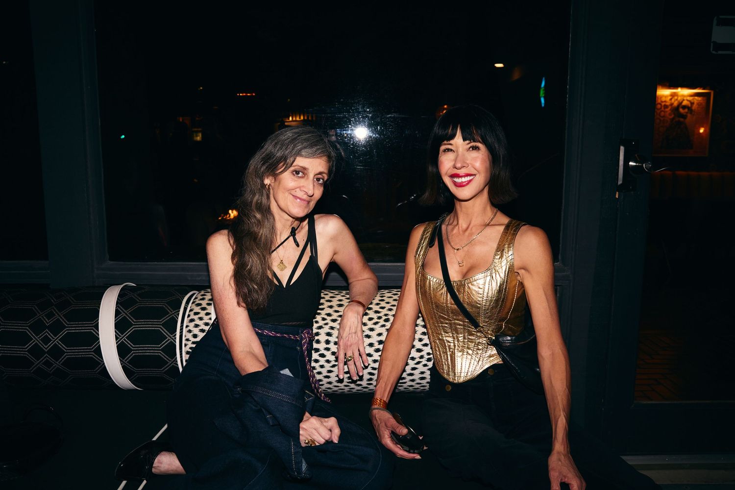 Two people in glamorous evening wear sit together at a dark venue with one wearing gold and one in black.