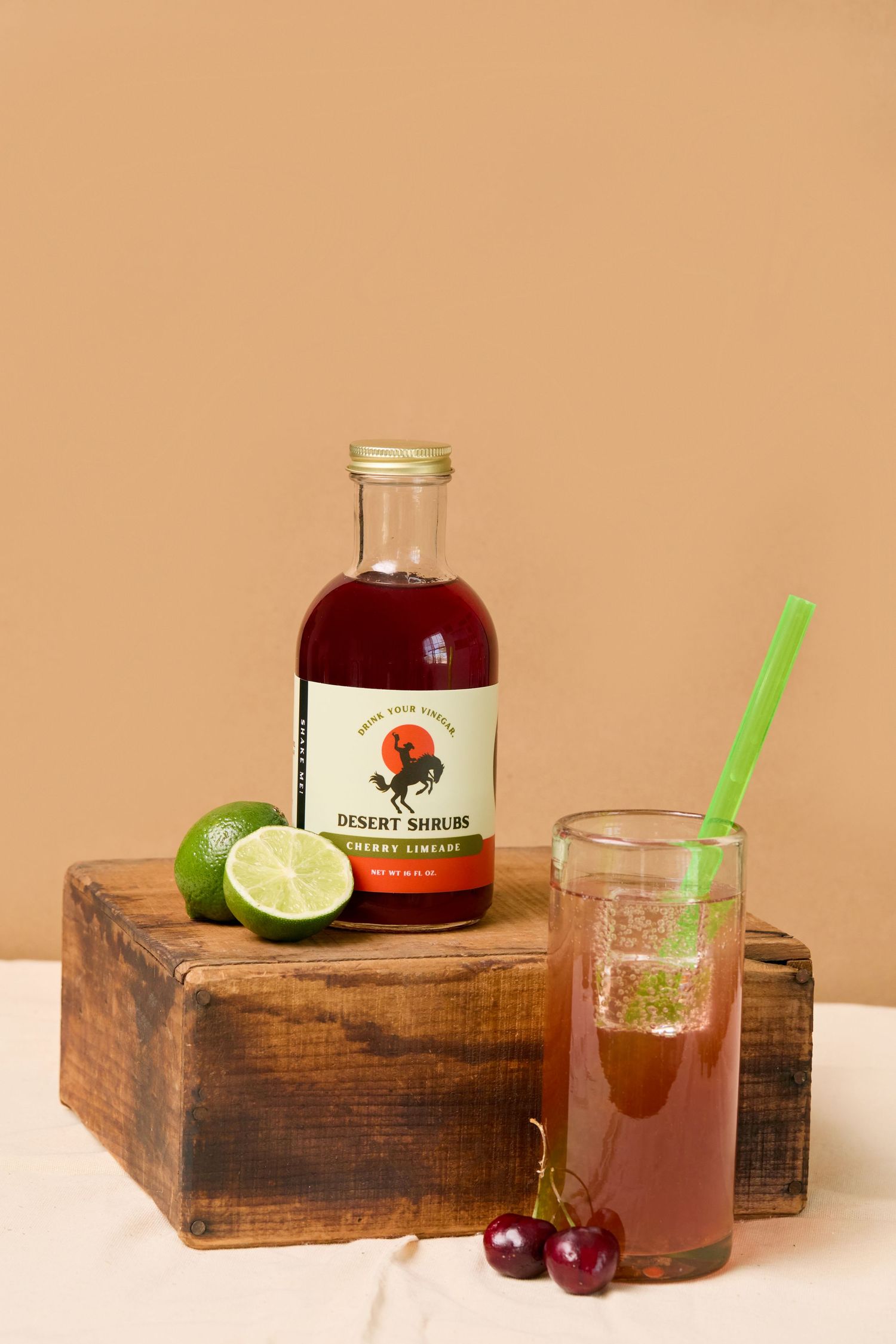 Desert Shrubs Cherry Limeade syrup bottle with fresh limes and a cherry cocktail drink with green straw.