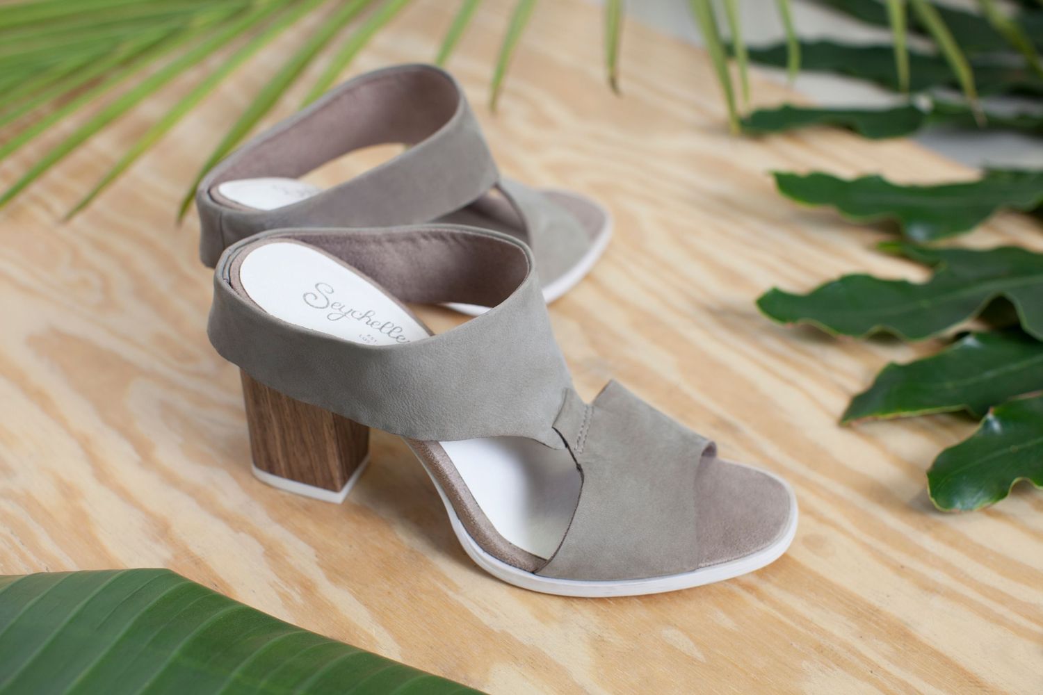 Modern gray suede sandal with block heel on wooden surface surrounded by tropical palm leaves.
