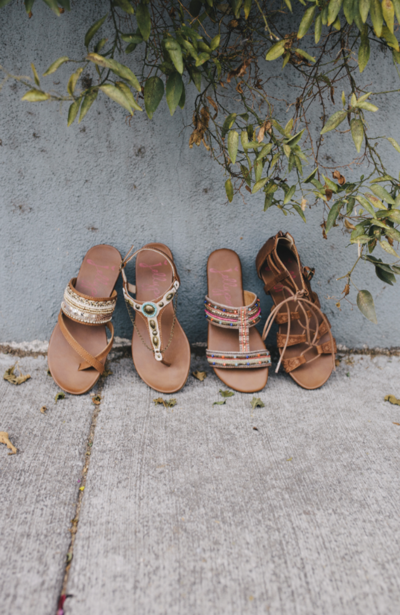 Four pairs of brown leather sandals lined up against a concrete wall with hanging foliage.