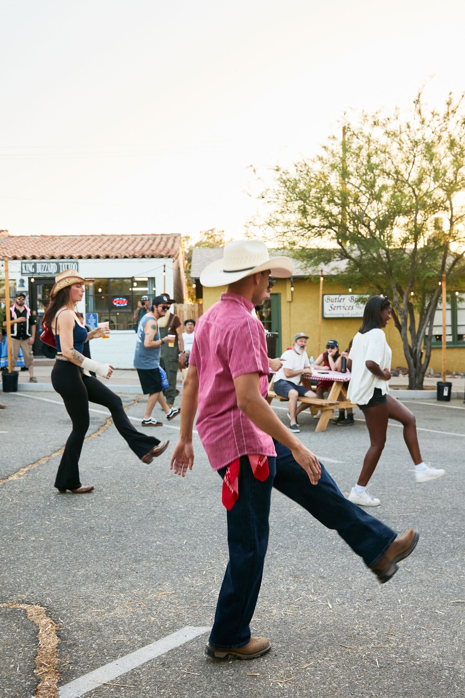 People line dancing outdoors in a parking lot, wearing cowboy hats and boots at a casual evening street event.