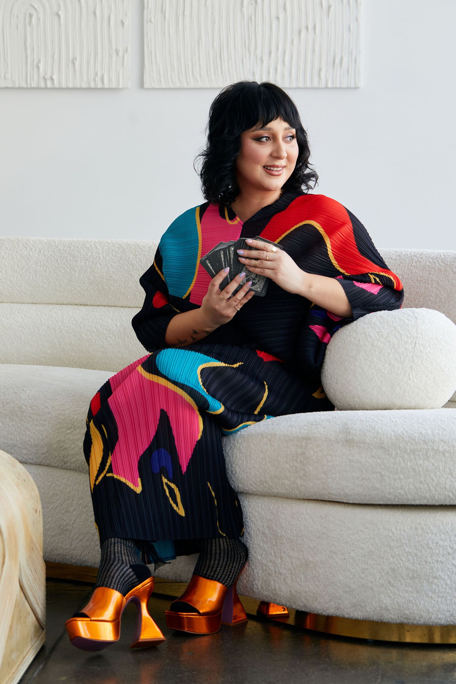 Woman in colorful printed outfit holding playing cards, seated on white sofa with orange platform heels.