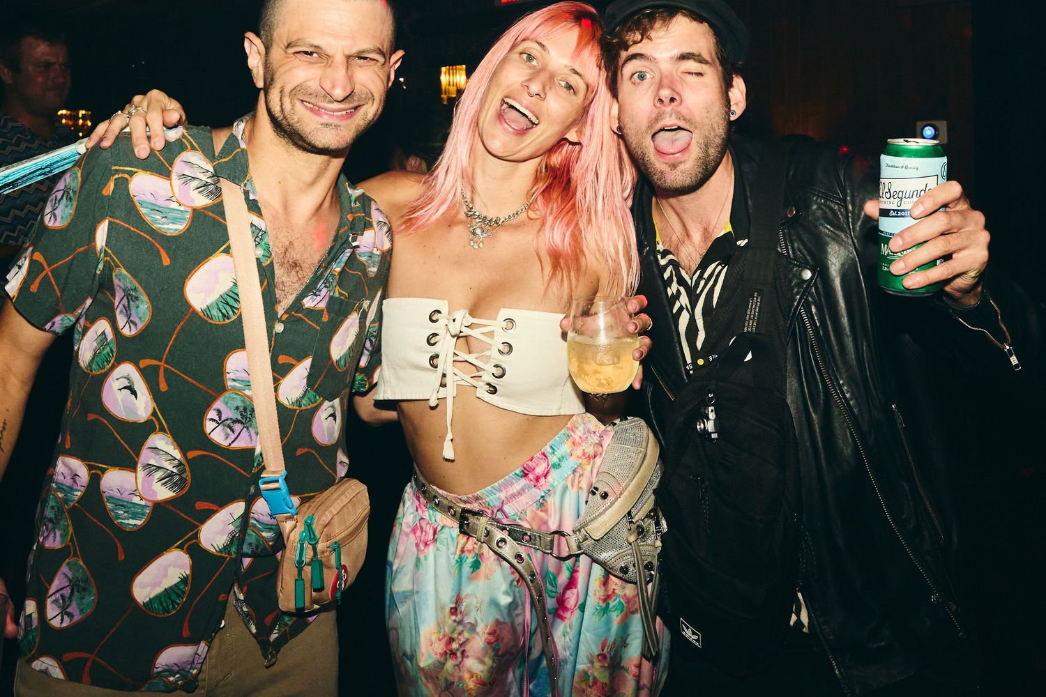 Three friends at a party enjoying themselves with drinks, one wearing a patterned shirt, one with pink hair, and one in leather.
