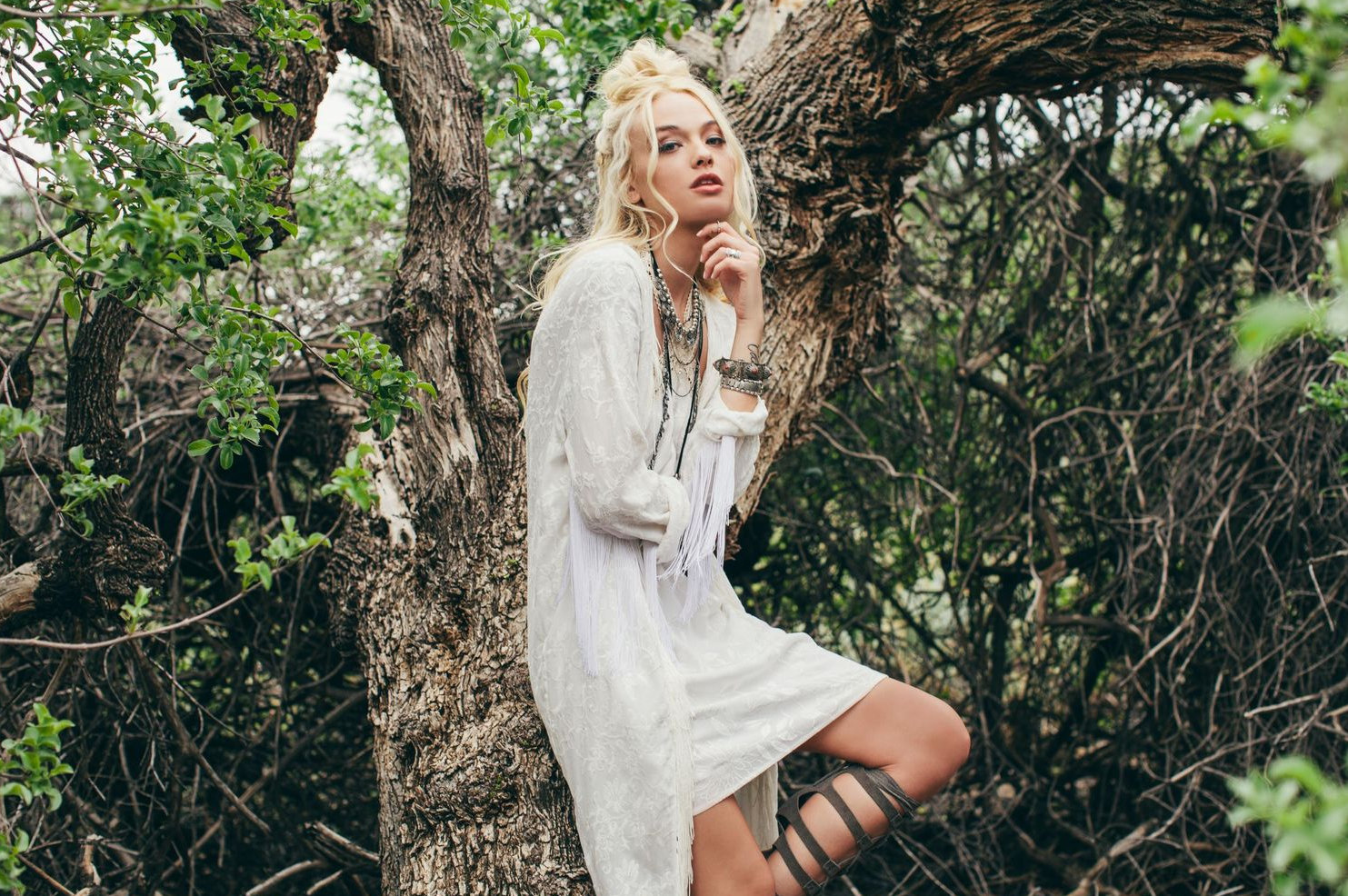 Model in white lace dress poses against tree in ethereal garden setting.