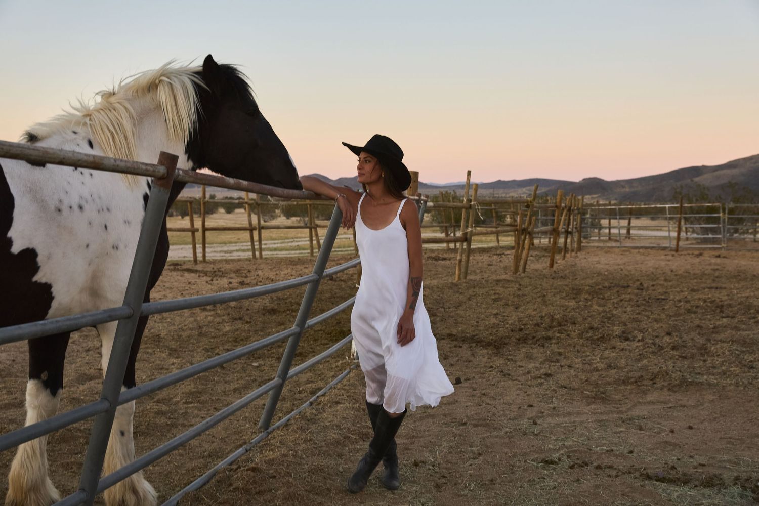 Woman in white dress and cowboy hat petting a black and white horse at a ranch fence during sunset.