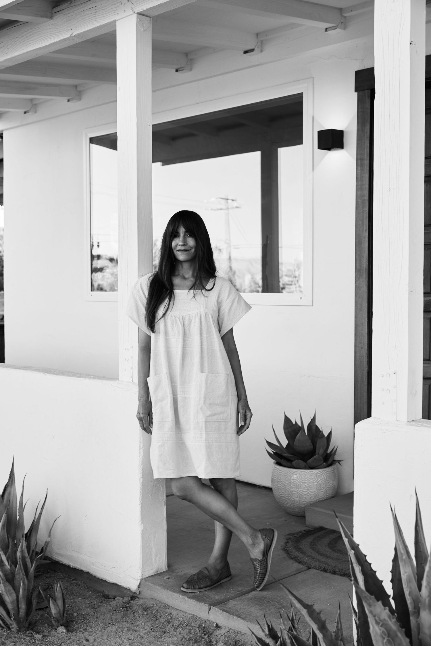 Black and white photo showing someone in a casual white dress standing on a porch with potted succulents and desert landscaping.