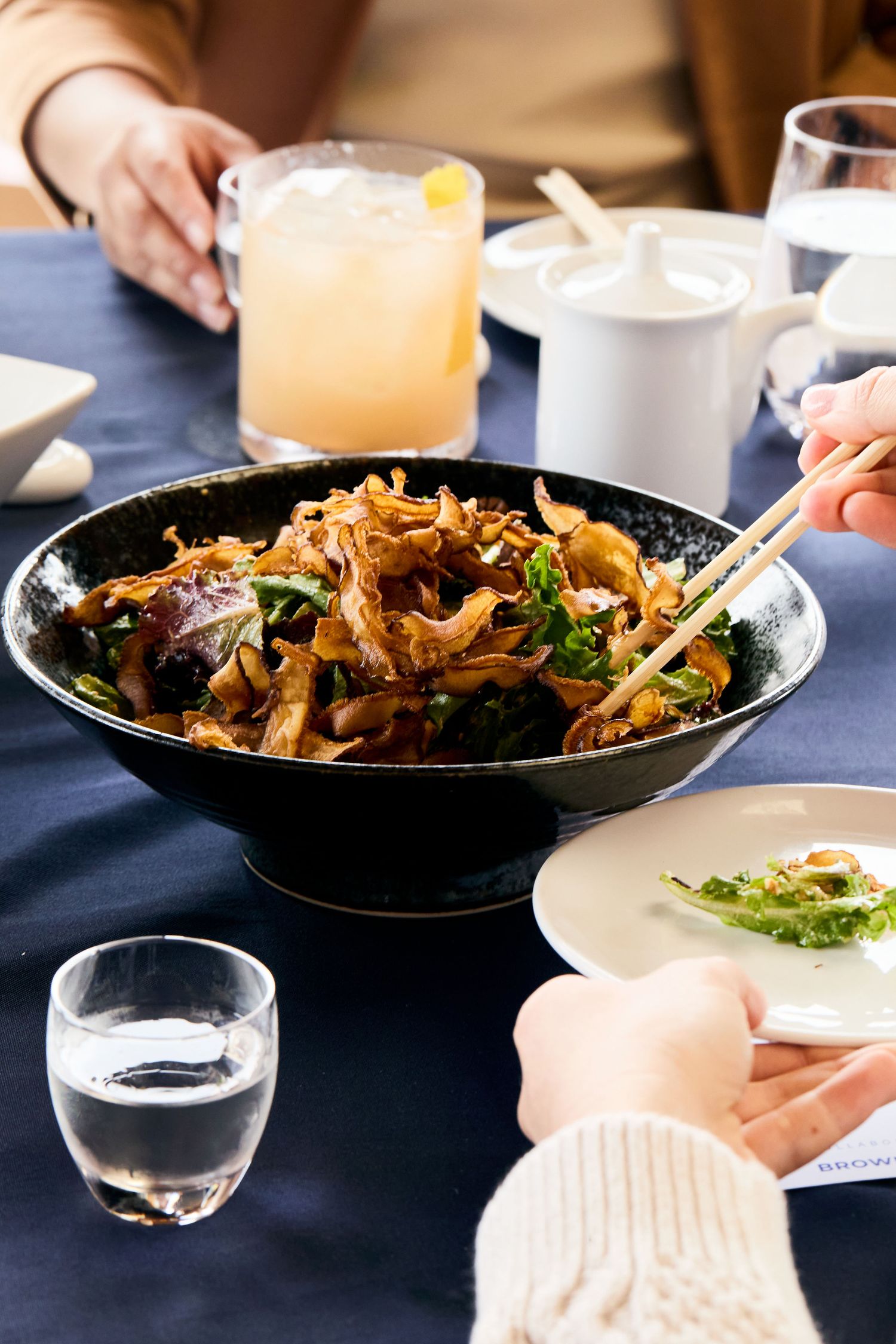 A bowl of stir-fried noodles with vegetables served alongside cocktails and water glasses on a blue tablecloth.