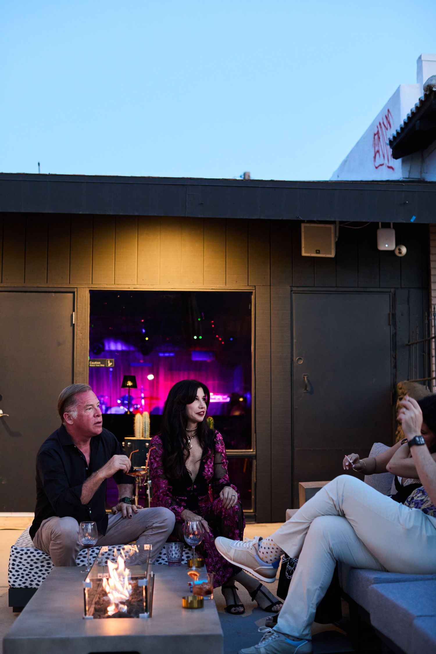 Friends relax on an outdoor patio at night with drinks and a candle centerpiece, purple lights glowing through doorway.