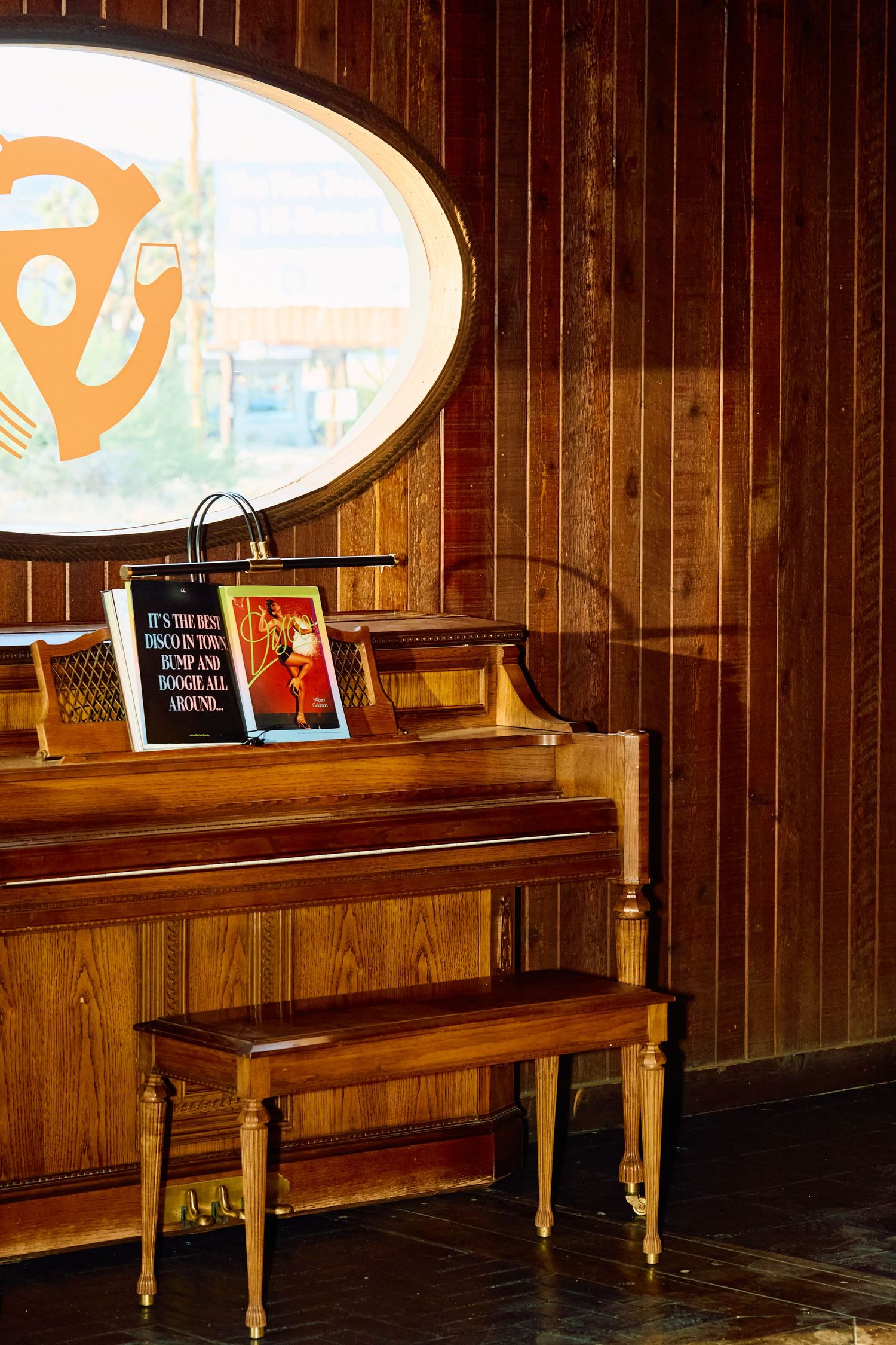 Vintage wooden piano and bench against wood-paneled wall with illuminated oval sign above in rustic interior setting.