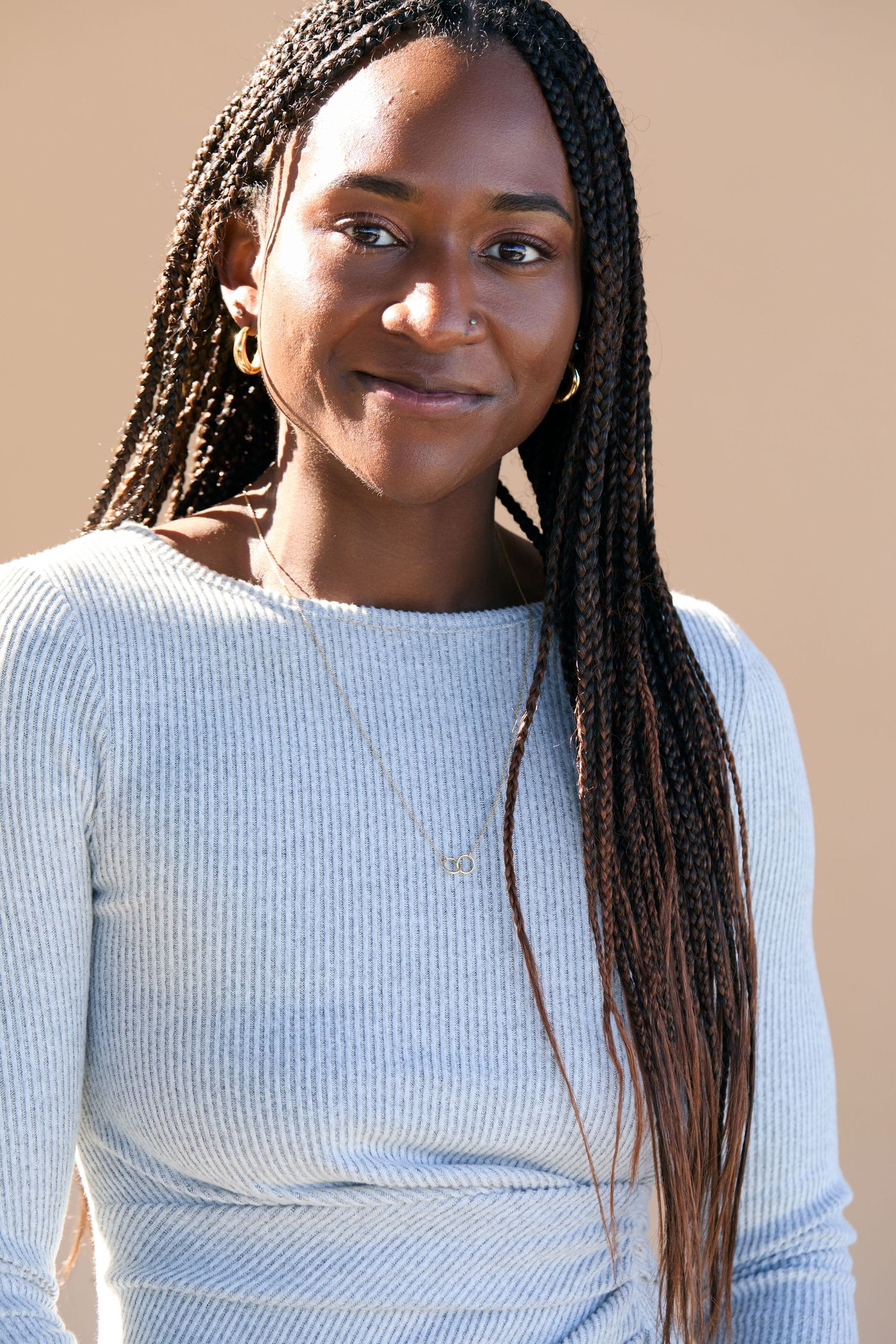 A person wearing a light gray ribbed sweater smiles warmly while showcasing long braided hair in natural lighting.